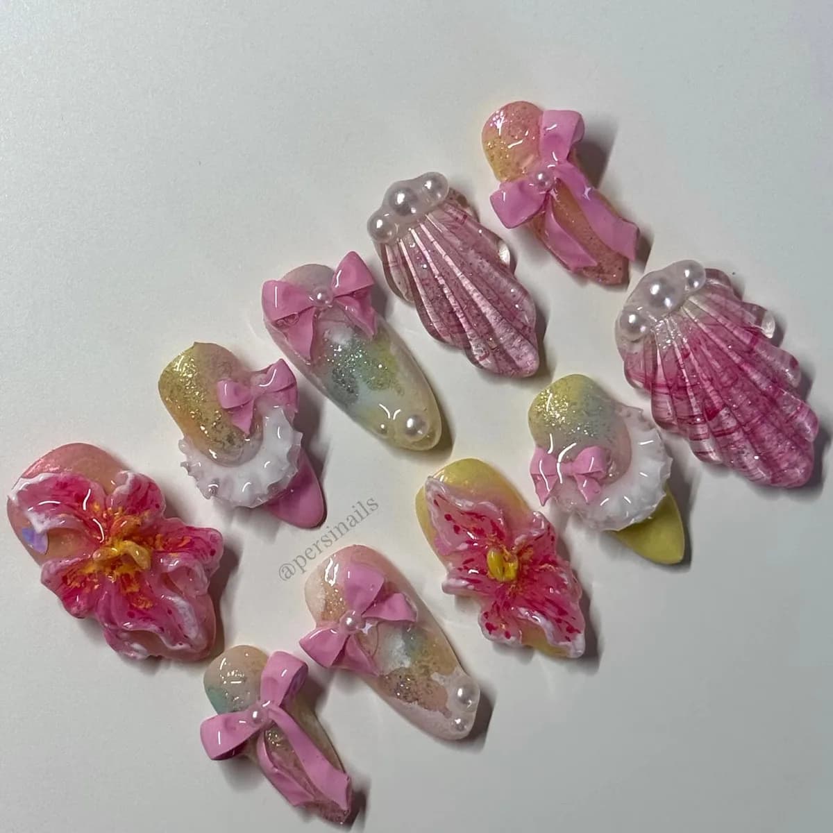 Ten press-on nails on a sheer iridescent base, with sculpted lilies, blush ribbon bows, ribbed shells and seed pearls.